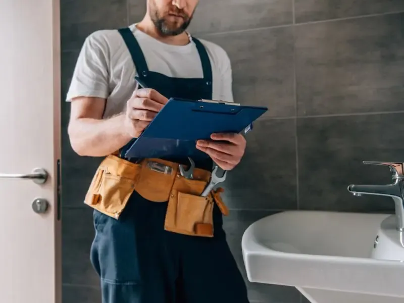 Un plombier prenant des notes sur un clipboard devant un lavabo dans une salle de bain.
